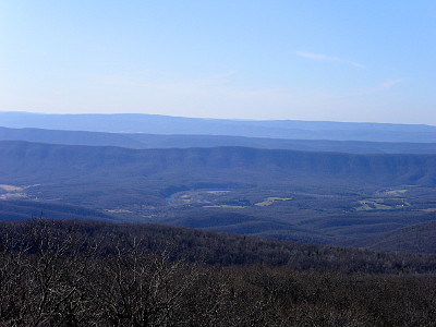 [This view from a high level looks down at a valley floor which had sections of trees removed although most remain. In the far distance are three levels of smooth-top mountain ridges all with varying levels, based on distance, of bluish-grey haze.]